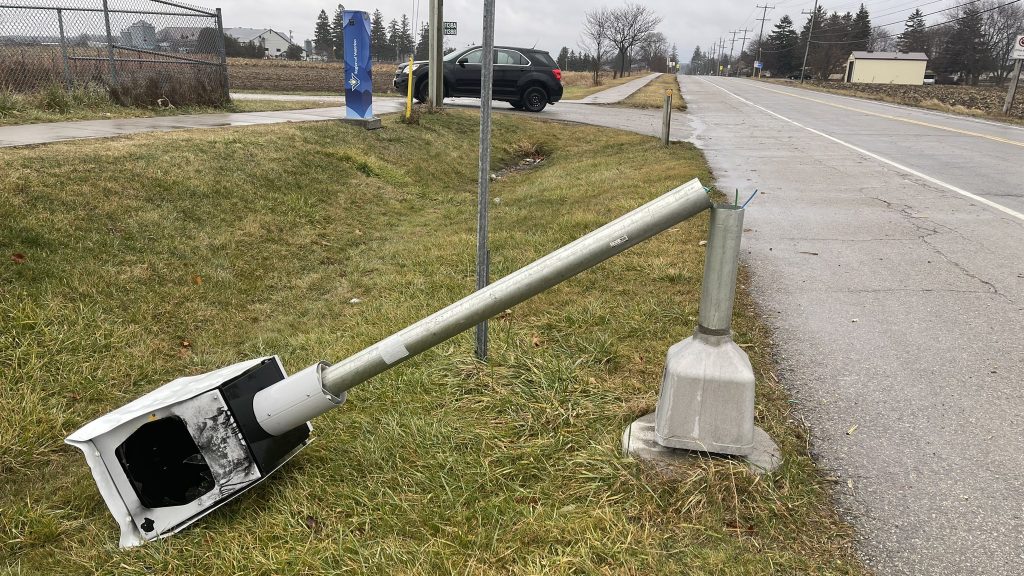 A grey Region of Waterloo traffic camera sits on the side of the road on a gloomy day, seemingly cut toward its base. Frayed wires are visible from the area where the traffic camera's support was cut. To the left side of the frame, the actual camera can be seen resting in the grass, its glass lens largely destroyed with only jagged shards visible.