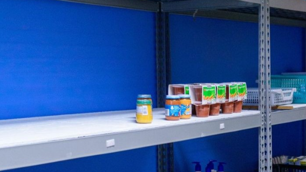 A metal shelf sits in front of a blue wall, scarcely populated with food items for donation, along with plastic miniature-hampers.