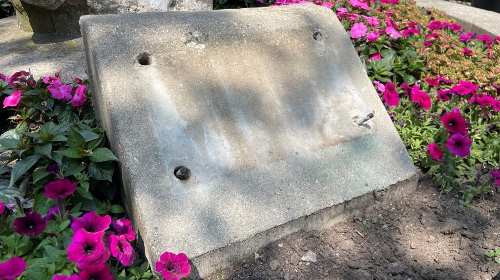 An empty stone placard sits in a bed of flowers, with water stains and four bolt holes where a plaque honouring a local police officer once was.