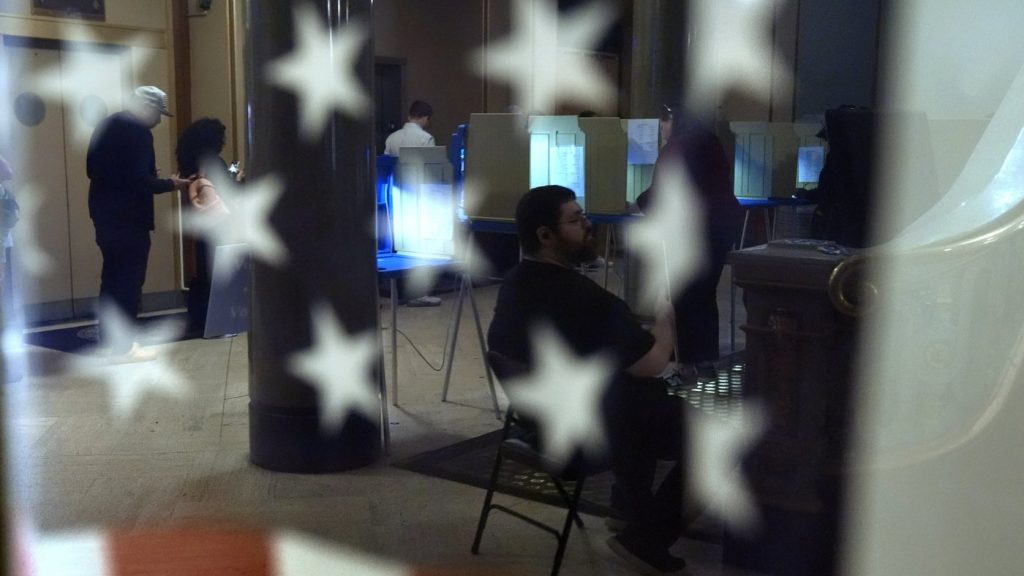 Voters are reflected in a window near an American flag.